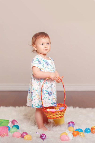 Cute adorable Caucasian baby girl in white dress celebrating traditional holy Christian Easter holiday. Kid child playing with colorful eggs in studio. Copyspace for text. - Photo, Image