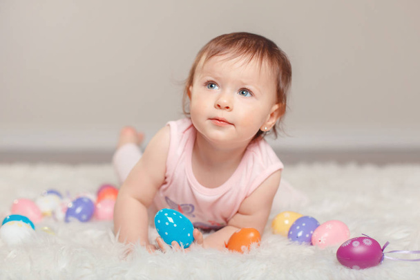 Cute adorable Caucasian baby girl in pink shirt lying on floor looking up and celebrating traditional Easter Christian holiday. Kid child holding and playing with colorful eggs - Foto, Imagen