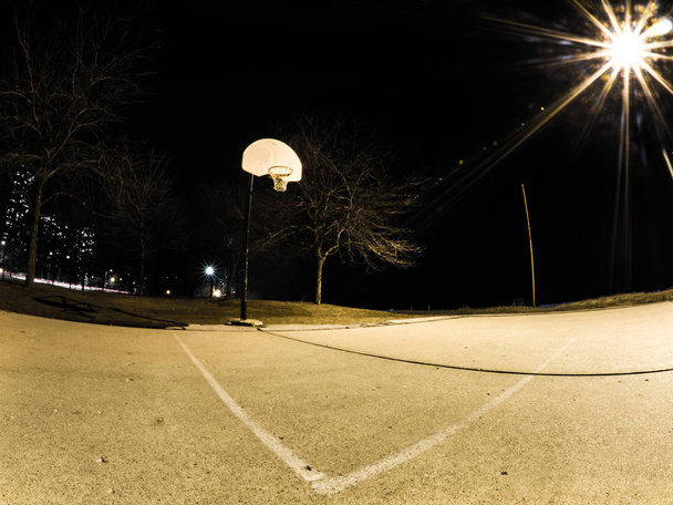 Ночная фотография открытой выветриваемой баскетбольной площадки и баскетбольных обручей с белыми носилками оранжевые диски и сети освещенные рядом уличным освещением в городе Чикаго
. - Фото, изображение