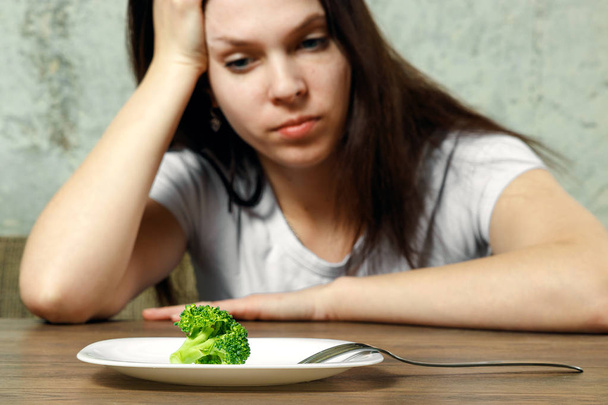 sad young brunette woman dealing with anorexia nervosa or bulimia having small green vegetable on plate. Dieting problems, eating disorder. - Fotografie, Obrázek