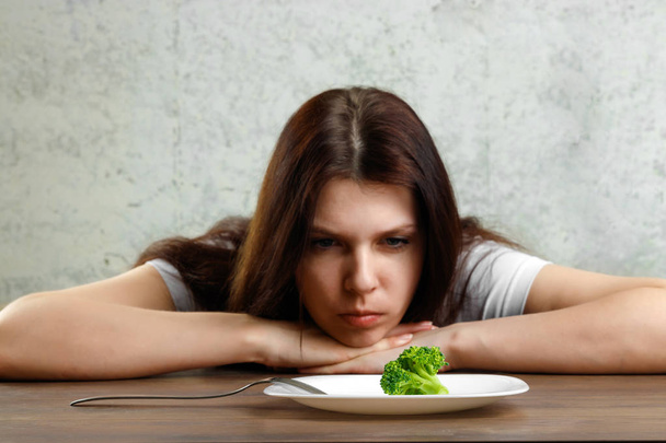 sad young brunette woman dealing with anorexia nervosa or bulimia having small green vegetable on plate. Dieting problems, eating disorder. - Foto, afbeelding