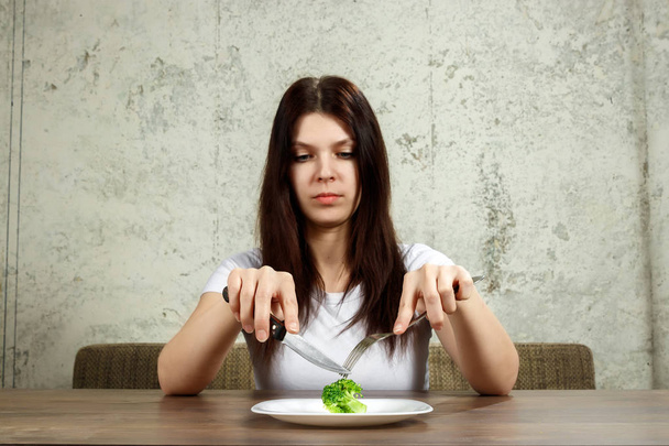 sad young brunette woman dealing with anorexia nervosa or bulimia having small green vegetable on plate. Dieting problems, eating disorder. - Zdjęcie, obraz