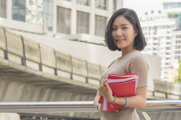 portrait of asian teen girl holds red book stands and dresses up in street fashion cloth at outdoor public space with modern building background - Photo, Image