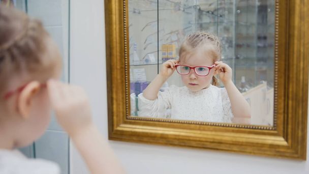 Маленькая девочка пробует новые очки возле зеркала - шоппинг в офтальмологической клинике
 - Фото, изображение