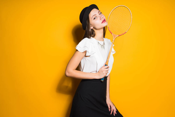 confident stylish girl model in fashionable black hat posing on yellow background, holding a tennis racket - Фото, зображення