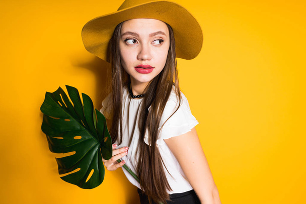 confiada hermosa chica en un sombrero de moda sostiene una hoja verde, posando sobre un fondo amarillo
 - Foto, Imagen