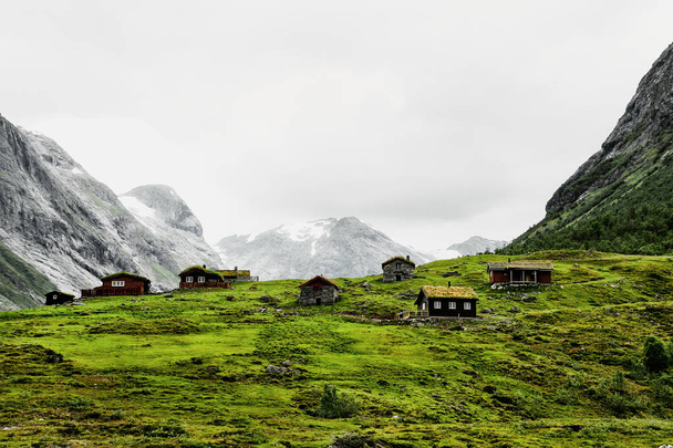 Pięknej przyrody na miejscu z rzeki, kamienie i pastwiska w dolinie pośród ośnieżonych szczytów górskich. Kolorowe domy stoją na zielonej trawie. Położony między Geiranger i Bergen w Norwegii. - Zdjęcie, obraz