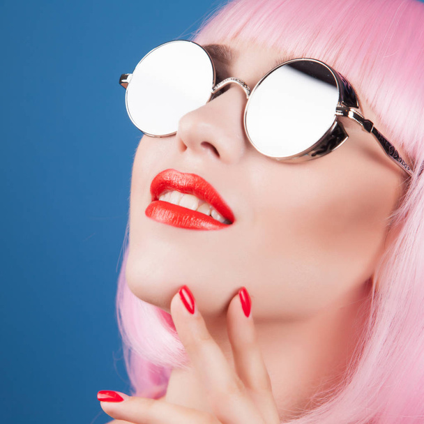 beautiful woman wearing colorful wig and silver sunglasses against blue background - Фото, изображение