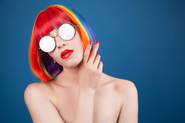 beautiful woman wearing colorful wig and silver sunglasses against blue background - Foto, Imagen