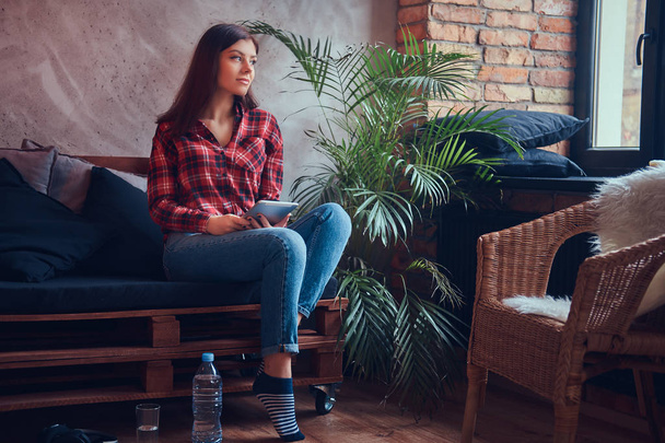 Morena encantadora em uma camisa de flanela e jeans sentados em um sofá
 - Foto, Imagem