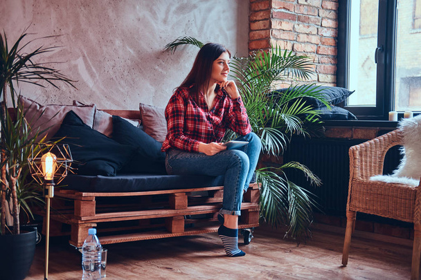 Charming brunette in a flannel shirt and jeans sitting on a couch - Zdjęcie, obraz