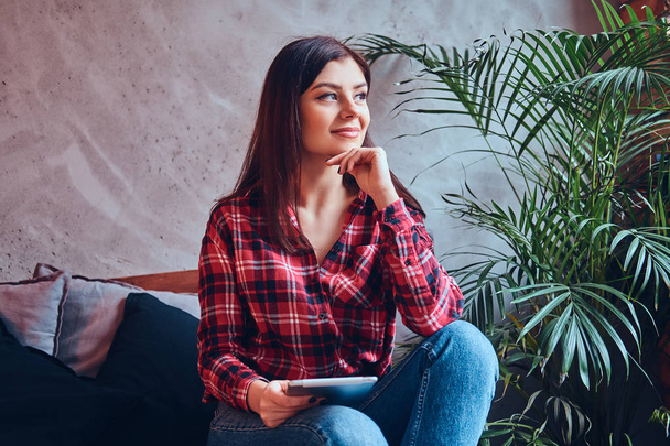 Charming brunette in a flannel shirt and jeans sitting on a couch - Foto, Bild