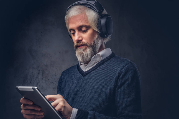 Portret przystojny starszego mężczyzny przy użyciu tabletu z słuchawki w studio na ciemnym tle. - Zdjęcie, obraz