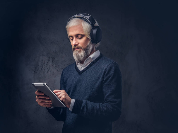 Portrait of a handsome senior man using a tablet with headphones in a studio over a dark background. - 写真・画像