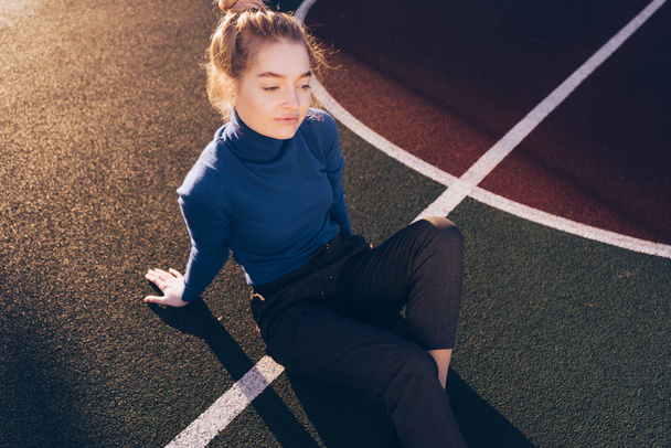 stylish beautiful young girl in a blue sweater sits on a sports field posing in the sun - 写真・画像
