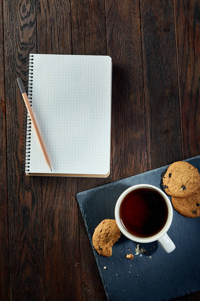 Φλιτζάνι τσάι με μπισκότα, το βιβλίο εργασίας και ένα μολύβι σε ένα ξύλινο υπόβαθρο, το top view - Φωτογραφία, εικόνα