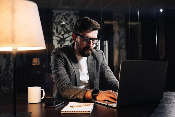Bearded young coworker typing text on modern laptop in loft office at night. Businessman working process. Man using contemporary notebook. Film effect - Fotoğraf, Görsel