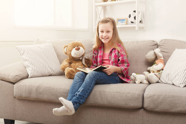Happy little female child hugging her teddy bear and reading book on sofa at home - Photo, Image