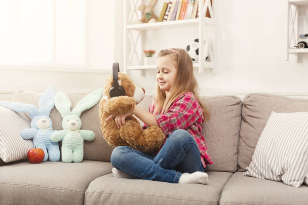 Boldog kis női gyerek és neki teddy bear zenehallgatás otthon a kanapén - Fotó, kép