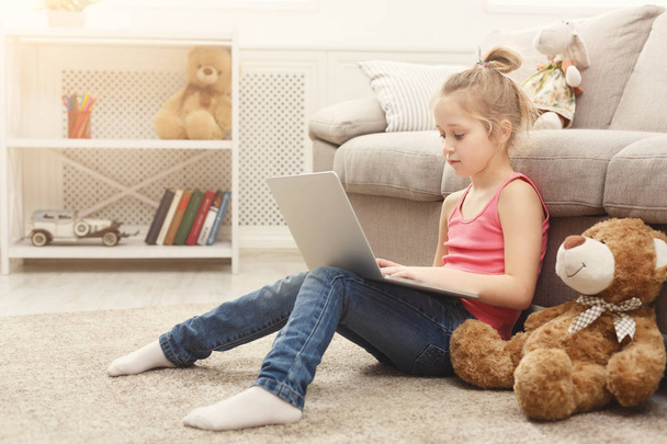 Schattig klein meisje huiswerk op laptop, zittend op de vloer thuis - Foto, afbeelding