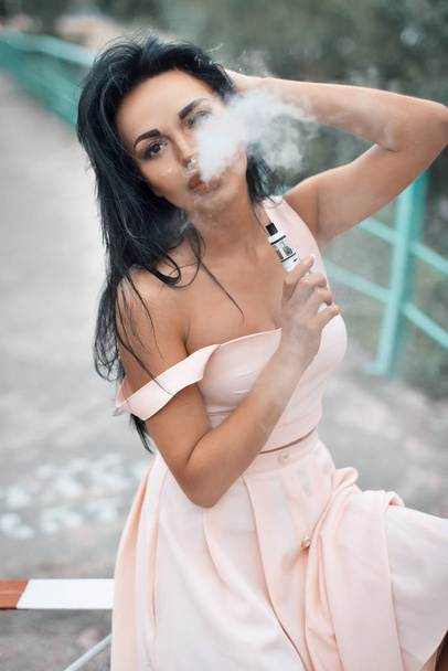 Young beautiful woman vaping e-cigarette with smoke outdoors. Vapor concept. - Foto, afbeelding