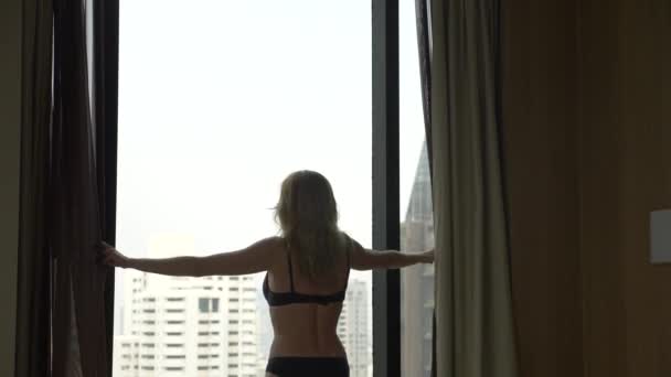 Een blonde vrouw in lingerie, opent de gordijnen op het venster weergeven vanuit het venster de wolkenkrabbers. Slow-motion. 4k - Video