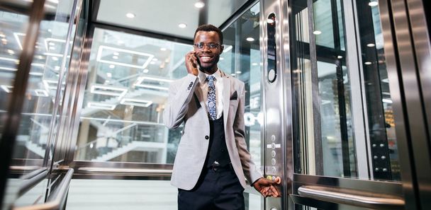 afroamerican businessman in modern glass elevator talking on phone - Fotó, kép