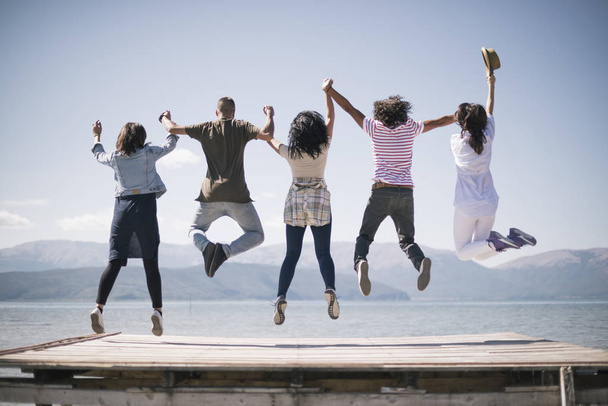 Group of happy young people jumping on a pier on a lake at a sunny day. View from back - Foto, afbeelding