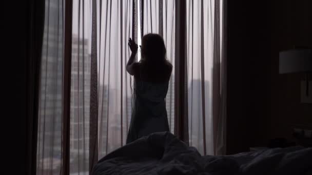 女性は、高層ビルを見渡せる窓に来て睡眠から目覚める、カーテンを開けます。スローモーション。4 k - 映像、動画