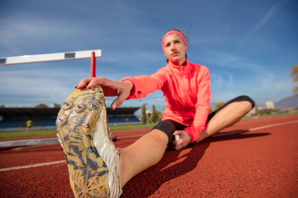Young sportswoman sitting and stretching on track field while listening to music, looking at camera. - Foto, Bild