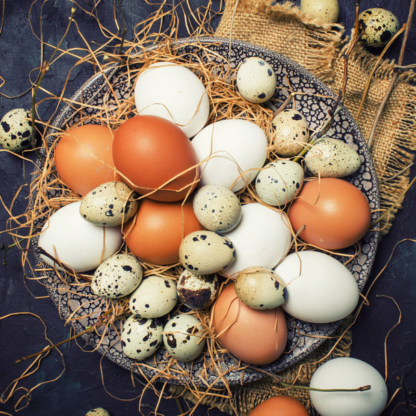Multicolored chicken and quail eggs with straw and branches, spring Easter composition, black background, top view - Foto, Bild