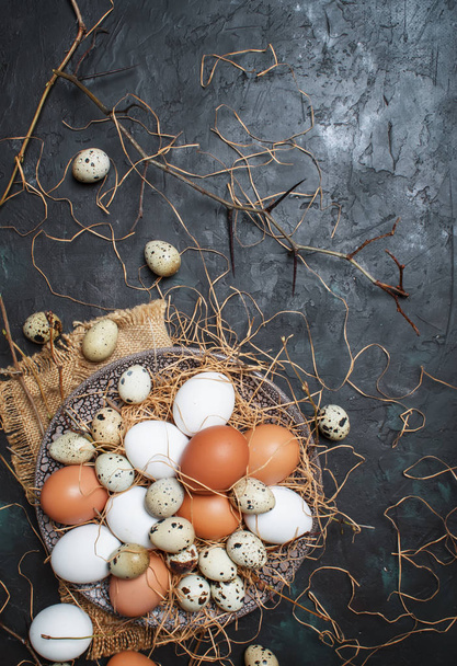 Multicolored chicken and quail eggs with straw and branches, spring Easter composition, black background, top view - Foto, afbeelding