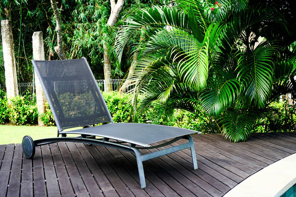 Une chaise longue se dresse sur un plancher de bois près de la piscine. En arrière-plan se trouvent des feuilles de palmier vert. Vue latérale avec espace de copie
 - Photo, image