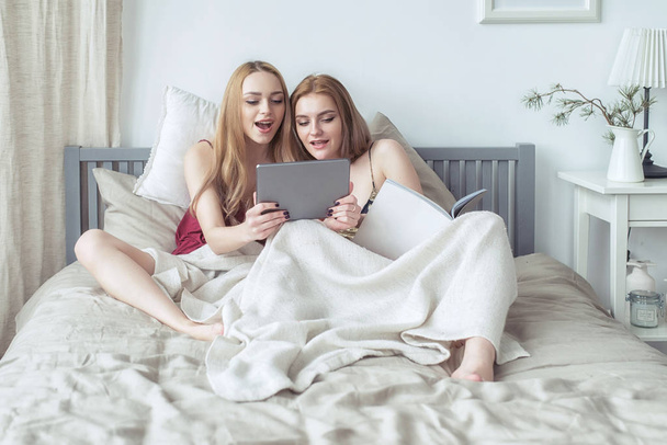 Two sexy blonde girls in pyjamas having fun in the bedroom. Young females lying in bed using tablet. - Foto, immagini