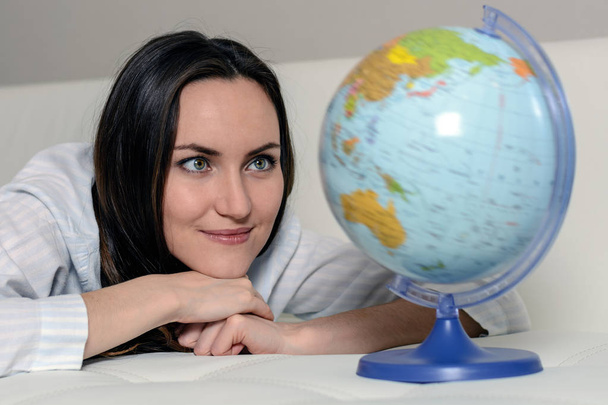 Закрытие - женщина в пижаме лежит на диване, глядя на земной шар со странами мира
 - Фото, изображение