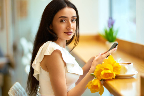 Beautiful Young Woman With Spring Tulips Flowers Bouquet At Cafe. Woman Drinking Coffee And Using Phone. Spring Style. Brunette Woman In Nice Spring.Beautiful Spring Mood. Fashion Spring/Summer. - Fotografie, Obrázek