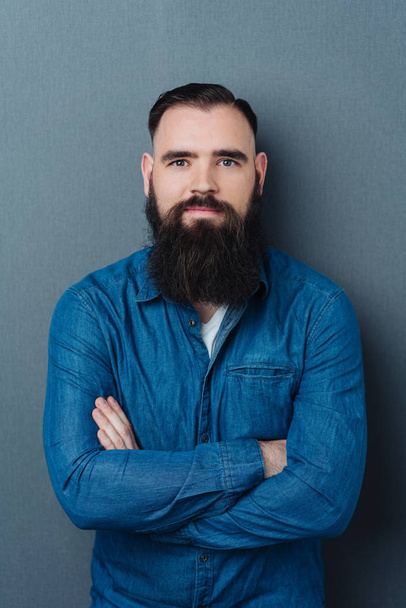 Confident bearded man in a casual blue denim shirt standing with folded arms over a dark grey studio background looking at the camera - Fotografie, Obrázek