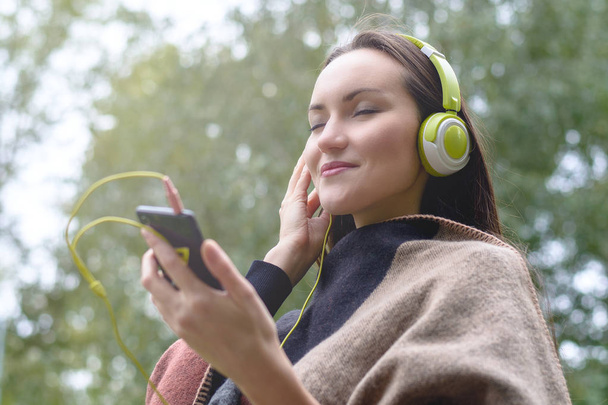 Młoda kobieta słuchania muzyki ze smartfona z słuchawek na świeżym powietrzu w parku. Widok z dołu - Zdjęcie, obraz