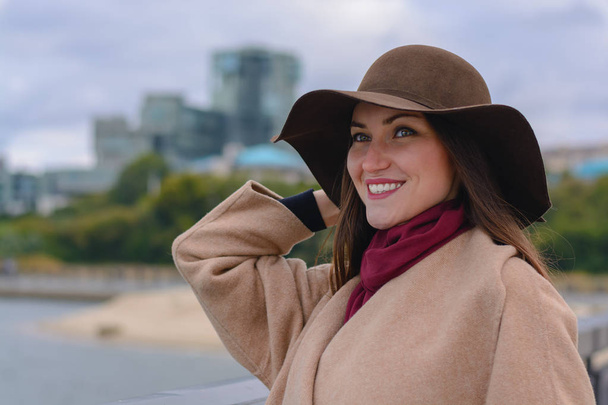 Retrato de una hermosa mujer joven de moda en un sombrero y abrigo en la orilla del río en la ciudad
. - Foto, Imagen