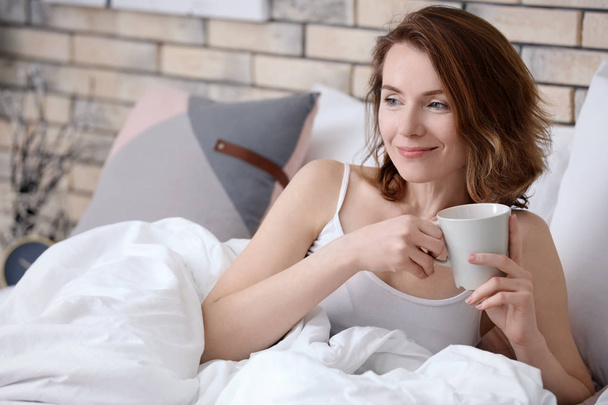 Beautiful smiling woman with cup of coffee lying in bed at home - Foto, afbeelding