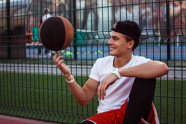 Oyun zamanı. Yakışıklı delikanlı turuncu topu tutarak ve basketbol sahası üzerinde dinlenme süre gülümseme ile kameraya seyir spor giyim - Fotoğraf, Görsel