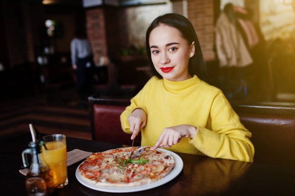 Funny brunette girl in yellow sweater eating pizza at restaurant - 写真・画像