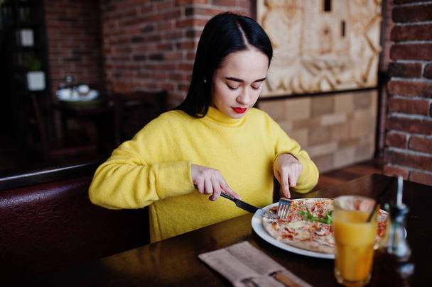 Funny brunette girl in yellow sweater eating pizza at restaurant - Zdjęcie, obraz