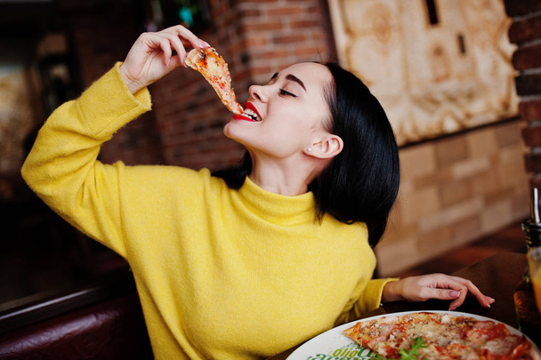 Funny brunette girl in yellow sweater eating pizza at restaurant - Foto, imagen