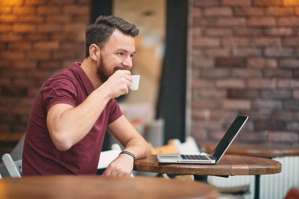 Homme buvant du café et utilisant un ordinateur portable tout en étant assis à la cafétéria
. - Photo, image