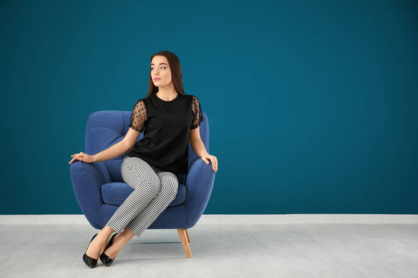 Beautiful young woman sitting in comfortable armchair against color wall - Valokuva, kuva