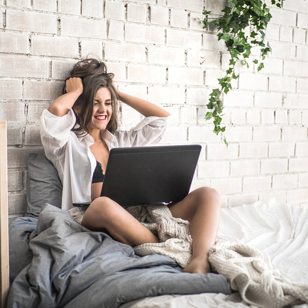 Eine aufgeregte junge schöne Frau mit erhobenen Armen sitzt im Bett und blickt auf ihren Laptop-Bildschirm. positive menschliche Emotion glückliche Mimik, Körpersprache, Reaktion. unerwartet angenehme gute Nachrichten - Foto, Bild