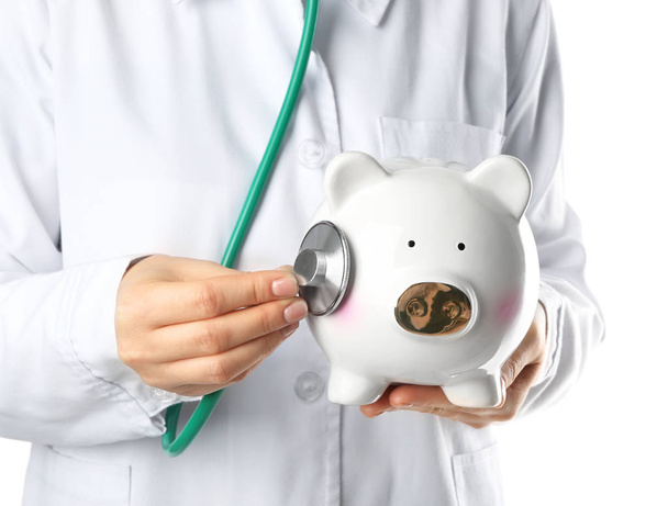 Young female doctor with ceramic piggy bank and stethoscope on white background - Foto, Bild