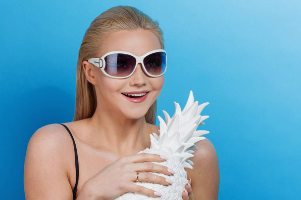 Young blond woman in blue bikini on blue background, with sunglasses and white pineapple - Fotoğraf, Görsel