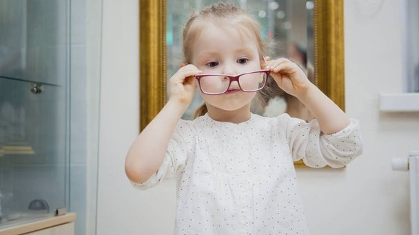 Little girl tries fashion medical glasses near mirror - shopping in ophthalmology clinic - Photo, image